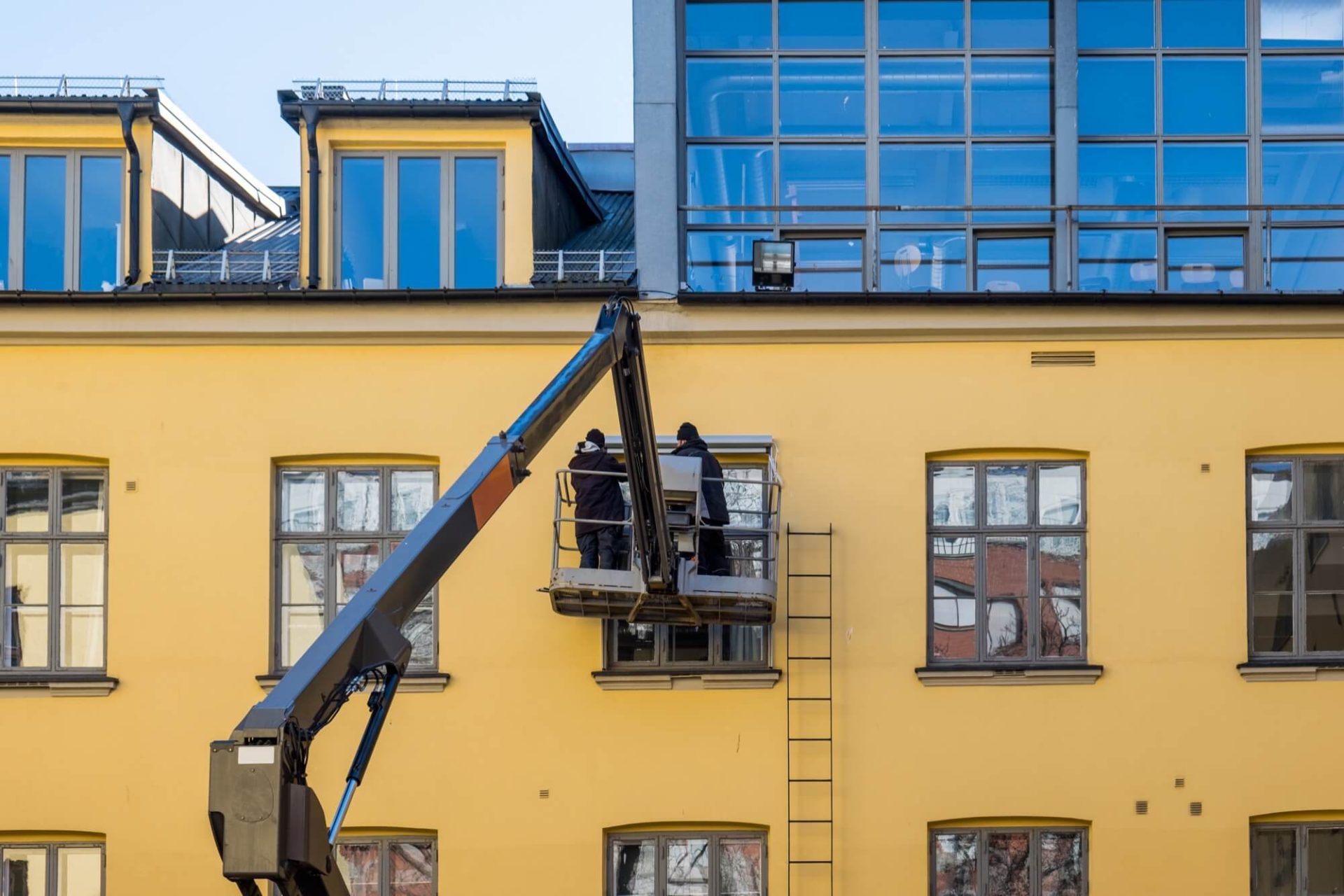 En gul byggnad får en ansiktslyftning, där man kan se två arbetare engagerade i fasadarbete från en lyftplattform